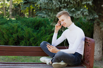 A young blond guy, dressed in a white jacket and black jeans, sits on a bench with headphones and looks at the phone against the background of the park. Life concept.