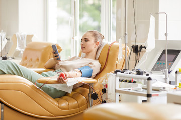 Young female donor lying on the couch and using her mobile phone while donating blood at hospital