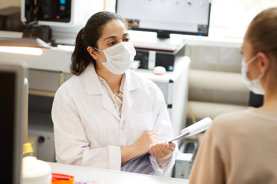 Female Doctor In Mask Making Notes In Medical Card While Talking To Patient At Hospital