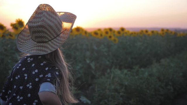 A happy little girl in the hat is smiling at the field of sunflowers at sunset time.