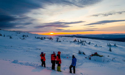 Four tourist are looking into the winter evening sky staying higher of the plato with trees. Sunset colors all around like winter tent, winter house, skis, equipment and many other things