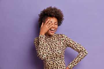 Carefree young Afro American woman covers face with palm, closes eyes and giggles positively, enjoys life, dressed in stylish leopard sweater, isolated on purple background, hears hilarious joke © WHstudio Leushin N