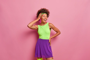 Joyful relaxed dark skinned woman expresses happiness, closes eyes with pleasure, smiles broadly, keeps hand on head, wears bright summer outfit, poses against pink background, being self confident