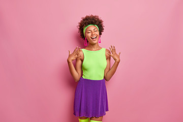 Happiness, pleasant moments of life. Overjoyed pleased woman with natural curly hair, closes eyes, laughs positively, raises hands, dressed in fasionable bright outfit, has fun, pink background
