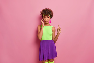 Serious thoughtful fashionable dark skinned woman talks via mobile phone, points index finger above, wears bright clothing, models against pink background. People, leisure, technology concept