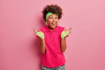 Studio shot of happy woman with natural curly hair, raises palms, feels confused, laughs positively, wears sport gloves, bright pink t shirt, poses indoor, cannot decide what is better for her