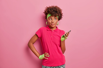 Sporty dark skinned curly haired woman keeps hand on waist, holds mobile phone in hand, uses special application for training online, wears headband, sport gloves, pink t shirt, stands indoor