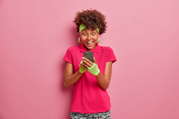 Excited happy woman stares with great happiness at smartphone display, being overemotional to read positive news, dressed in casual t shirt and headband, rosy background, receieves money on account