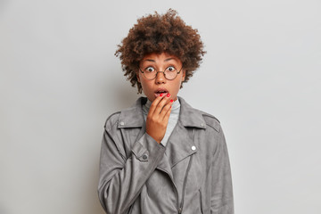 Amazed speecless woman covers mouth, stares in disbelief, shocked to realize her mistake, wears round spectacles, grey jacket, stands isolated. Human face expressions, emotions, reaction concept