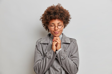 Fototapeta na wymiar Calm hopeful young woman with curly hair, keeps hands together, prays and hopes for better, enjoys peaceful atmosphere, wears spectacles and grey jacket, anticipates something very important