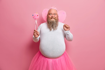 Positive plump man has fun on theme birthday party, feels like fairy who makes dreams come true, chills with children, has thick beard and fat belly, poses with magic wand and wings on back.