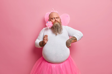 Fototapeta premium Serious bearded man thinks how to entertain children on party, wears funny costume of princess or fairy, concentrated thoughtfully aside. Family, celebration, fatherhood, entertainment concept