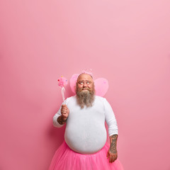 Funny plump daddy plays fairy on birthday party, amuses children, foolishes around, looks hopefully above as makes wish with magic wand, pink background. Man acts out fairytale has fun plays with kids