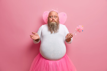 Big bearded man fairy with wings looks confused, spreads palms, organises costume party, poses with...