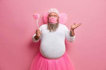 Fototapeta premium Online upbringing during quarantine. Shocked man wears fairy clothes, pink mask, holds magic wand, tries to entertain child via internet, stands indoor against rosy wall, stays at home, feels hesitant