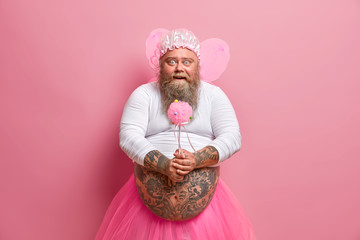Horizontal shot of bearded fatso man foolishes around on childrens birthday party, holds magic wand wears fairy costume, has fat tattooed stomach, isolated over pink background. Happy festival