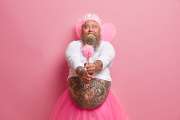 Male performer or holiday animator works on party for little children, has funny expression, holds magic wand, dressed like fairy, isolated on pink background, wears fancy wings. Man from wonderland
