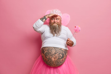 Photo of funny bearded father dressed in carnival costume, plays with children, wears fairy wings, undersized jumper and rosy pleated skirt, holds magic wand. Celebration, holiday, masquerade concept