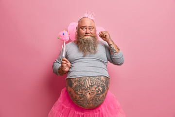Handsome positive fat man curls mustache, has fun on party, feels like real princess, wears crown, wings, organises baby shower for her newborn daughter. Beared fatso fairy has childish mood
