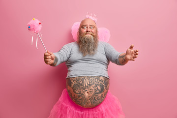 Studio shot of friendly looking kind fatso man wears kids costume for new year party, dressed in fairy outfit, spreads hands and wants to hug someone, isolated on pink background. Happy entertainment - Powered by Adobe