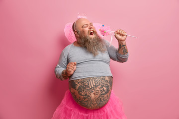 Overjoyed bearded plump man wears fairy clothes, holds magic wand near mouth as microphone, sings songs, foolishes around indoor. Dad plays with kids, acts like princess, isolated on pink background