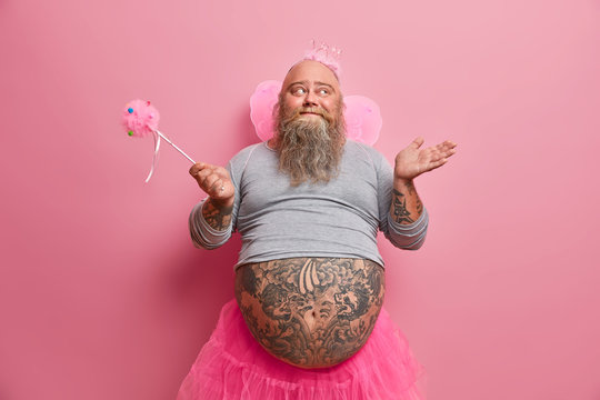 Happy kind man has image of innocent princess, wears crown, butterfly wings, holds magic wand, spends free time on costume party, poses indoor. Funny overweight male fairy plays with kids at home