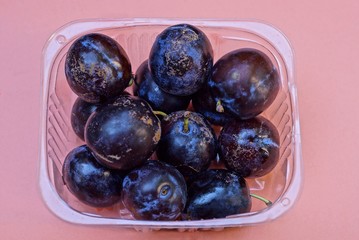 ripe blue plums in a transparent plastic box stands on a pink table