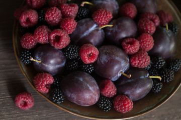 Raspberries, blackberries, plums in a bowl on a wooden background. Fragment, blur, selective focus.
