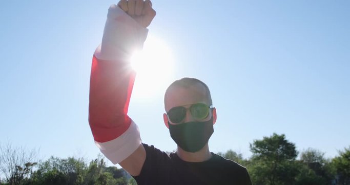 A man in a black mask and sunglasses raised up his hand with the coiled on it flag of Belarus against the backdrop of the back sun and green trees.