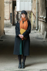 Long-haired girl student in a black coat with a vintage cap and a red scarf, posing with a book in a shabby quiet courtyard