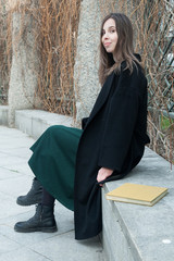 Brunette long haired girl sitting on a bench in a spring park near hedge without leaves