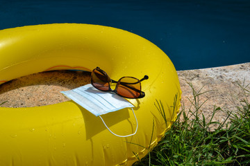 Blue face mask for protection against the Corona Virus (COVID-19), sunglasses laying on a pool mat beside a swimming pool during coronavirus worldwide pandemic holidays
