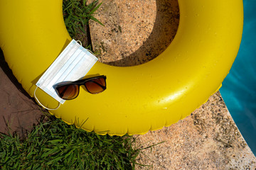 Blue face mask for protection against the Corona Virus (COVID-19), sunglasses laying on a pool mat beside a swimming pool during coronavirus worldwide pandemic holidays