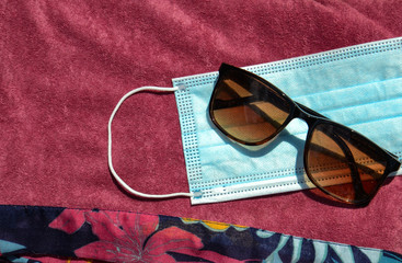 Blue face mask for protection against the Corona Virus (COVID-19), sunglasses laying beside a swimming pool during coronavirus worldwide pandemic holidays