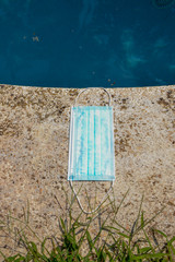 Blue face mask for protection against the Corona Virus (COVID-19), laying beside a swimming pool during coronavirus worldwide pandemic holidays