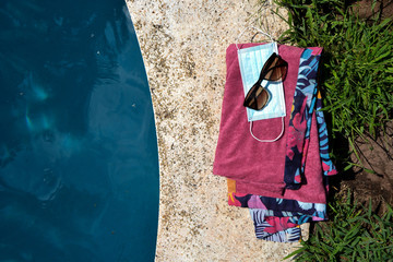 Blue face mask for protection against the Corona Virus (COVID-19), sunglasses and a pink towel laying beside a swimming pool during coronavirus worldwide pandemic holidays