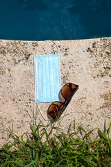 Blue face mask for protection against the Corona Virus (COVID-19), sunglasses laying beside a swimming pool during coronavirus worldwide pandemic holidays