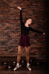 Beautiful young female classical ballet dancer on pointe shoes wearing a black leotard and skirt on a brick background