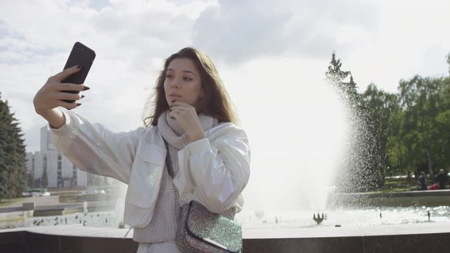 A side view slow motion close up shot of a dark-haired woman in white clothes standing in front of the fountain and taking pictures of herself