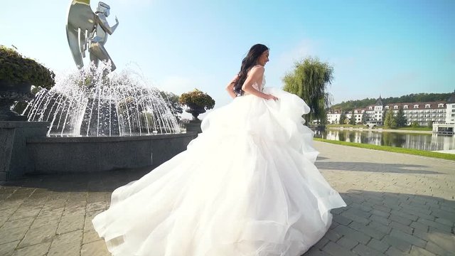 fashion bride runs and looks around at the camera. Young brunette woman in a luxurious wedding dress runs away One near the fountain on a sunny day with long train. Slow motion Rear full-length view