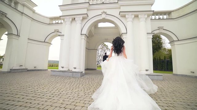 bride in a wedding dress runs to groom at sunset or sunrise against the backdrop of beautiful architecture. Slow motion. Europe. Fashion young woman goes to her husband hugs him and kisses Slow motion