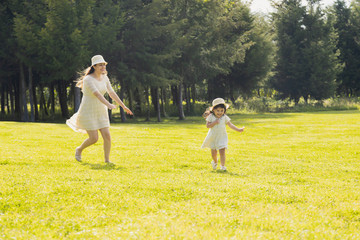 Naklejka na ściany i meble Mother and daughter playing and running in a green field with trees in the afternoon - mother and daughter wearing white dresses in the middle of a natural park