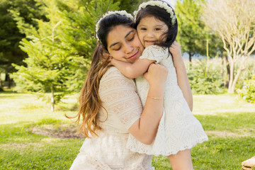 Hispanic mom and daughter hugging in a natural park surrounded by trees - mom and daughter dressed in white in the field