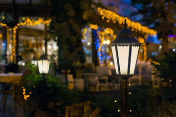 iron retro lantern of cafe lighting glowe with warm light in the night garden of the backyard in the background bokeh balls from garland on the facade restaurant building, nobody.