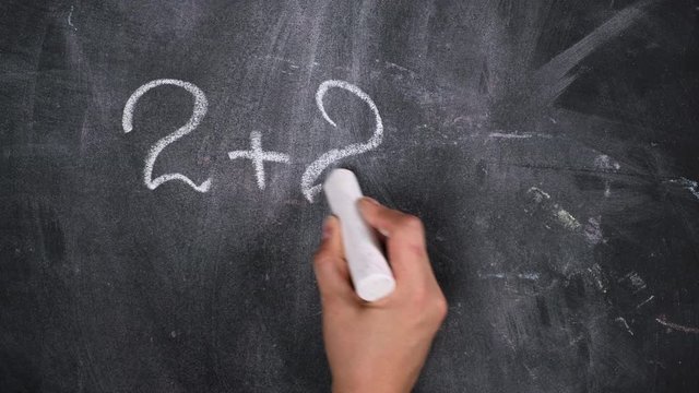 math example written in white chalk on a black chalk board and a woman's hand with white chalk, two plus two