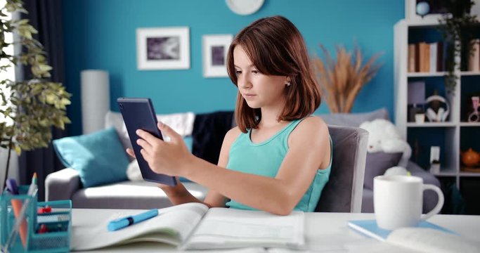Cute female teenager sitting at desk and using digital tablet. Pretty girl with brown hair studying at home on modern gadget. Concept of people, technology and education.