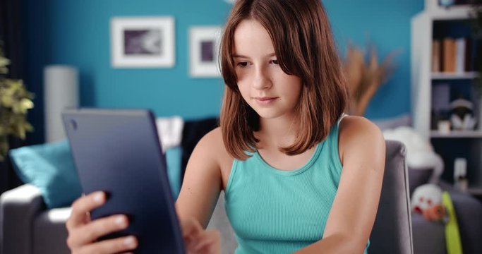 Positive teen girl sitting at desk and using digital tablet. Beautiful adolescent in casual outfit spending free time at home with modern technologies.