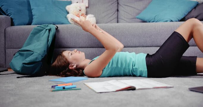Pretty female adolescent in casual clothing reading new book while lying on floor at own room. Young girl with dark hair enjoying free time with interesting literature.
