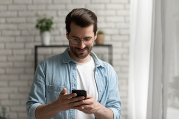 Obraz na płótnie Canvas Happy millennial caucasian man in glasses look at smartphone screen browse wireless internet on gadget, smiling young male text message on modern cellphone at home, communication concept