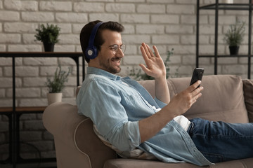 Smiling millennial Caucasian man in headphones wave greeting talk on video call on smartphone,...